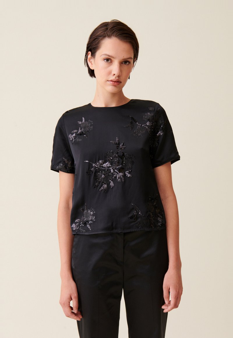 Blusa nera in satin a maniche corte con ricami floreali e paillettes. Collo rotondo, vestibilità morbida, abbinata a pantaloni neri coordinati.