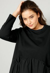 Černé šaty s dlouhými rukávy, kulatým výstřihem a nařasenou sukní se dvěma bočními kapsami. Hladká látka s volným střihem.