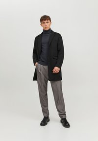 Manteau long noir sur un col roulé gris, associé à un pantalon à rayures fines gris clair et des chaussures noires à lacets, présenté sur un fond neutre.