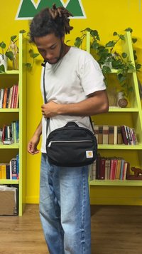 Crna najlonska torba preko tijela s prednjim džepom na patentni zatvarač i logotipom brenda. Nosi je osoba, ima podesivu naramenicu i zaobljeni oblik.