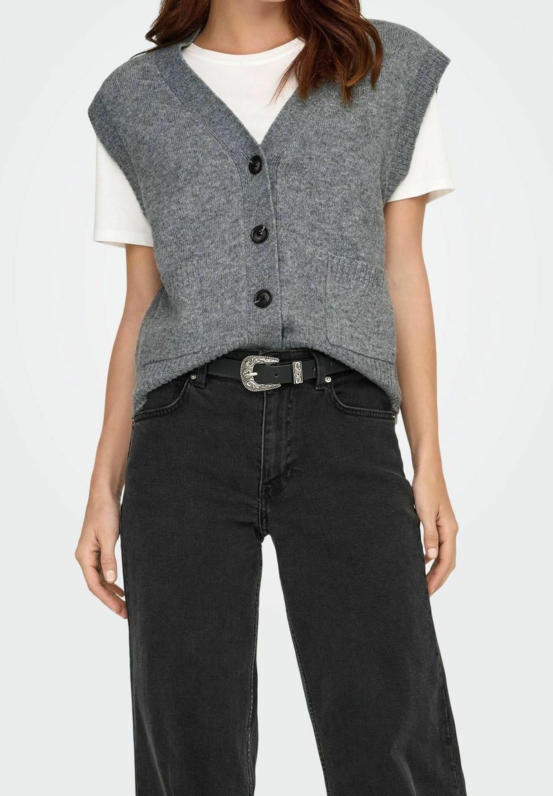 Femme portant un gilet gris tricoté sans manches boutonné sur une chemise blanche à manches courtes, un jean noir taille haute et une ceinture noire.