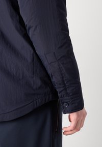 Chaqueta ligera azul marino que presenta una textura suave, puños abotonados y un detalle de cremallera en el dobladillo, con un diseño minimalista.
