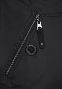 Detalj av svart jacka med en silverfärgad dragkedja, svart metalltryckknapp och texturerat tyg med sömnadsaccent.