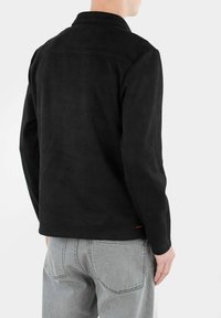Giacca in suede nera con collo a camicia, dettaglio sullo yoke posteriore e cuciture minimali. Abbinata a jeans grigi chiari.