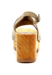 Zoccolo in suede beige con tacco in legno. Presenta una cinghia alla caviglia sul retro e dettagli metallici decorativi. La superficie ha una texture liscia.