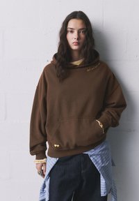 Hnedá oversized mikina s predným vreckom, zdobená žltým vyšívaným textom a srdiečkami na leme. Nosená s viacerými tričkami pod ňou.