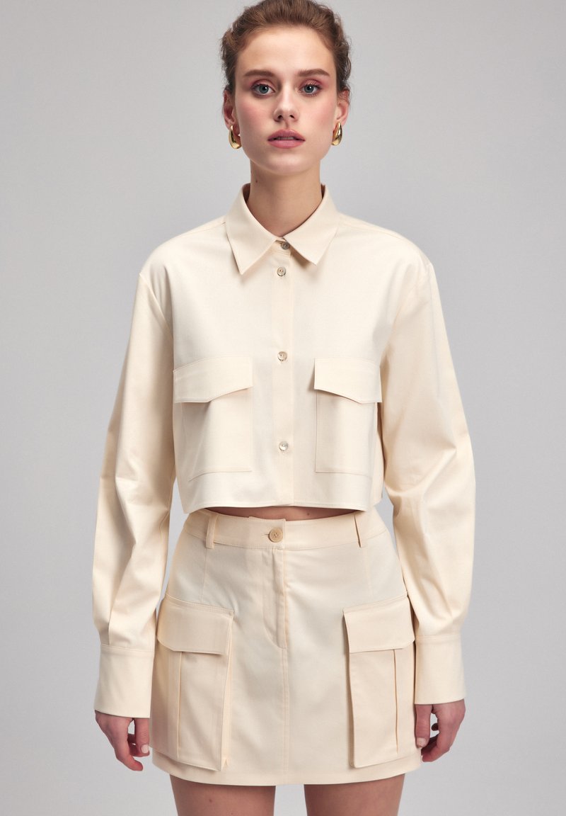 Kort beige skjorta med två stora bröstfickor och knäppning, matchad med en kjol i samma tyg med fickor, båda i ett mjukt material.