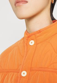 Oranžová prošívaná látková bunda s vysokým límcem, krémovými knoflíky a jemně vyšitými vzory pro texturu a designové detaily.