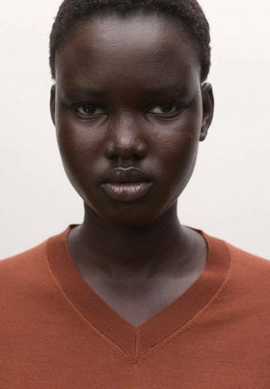 Primer plano de una persona con cabello corto y rizado, que lleva un suéter de cuello en V color óxido, mirando directamente a la cámara con una expresión neutral.