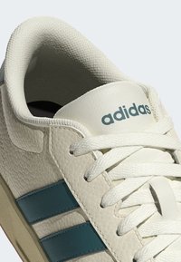 Bijela kožna tenisica s tirkiznim prugama, teksturiranim gornjim dijelom i bijelim vezicama. "adidas" logo otisnut na jeziku. Zaokružen vrh, gumeni potplat.