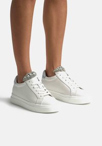 Sneakers bianchi in pelle con un design elegante, punta rotonda e rifiniture argento, arricchiti da un colletto decorato di gioielli attorno alla caviglia. Suola piatta.