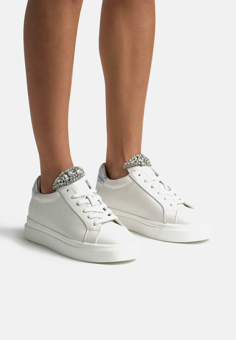 Sneakers bianchi in pelle con un design elegante, punta rotonda e rifiniture argento, arricchiti da un colletto decorato di gioielli attorno alla caviglia. Suola piatta.