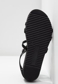 Semelle noire de sandale avec de profondes rainures horizontales et le nom de la marque "Tamaris" embossé au centre, appuyée contre une surface blanche.