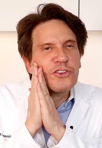 Manlig figur i vit laboratoriefrack, med ljust brunt hår, händerna på kinderna, blårandig skjorta synlig under, neutral bakgrund.