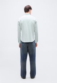 Άνδρας όρθιος με πλάτη φορώντας ανοιχτό πράσινο πουκάμισο με μακριά μανίκια και άνετο μπλε τζιν, μπροστά σε απλό λευκό φόντο.