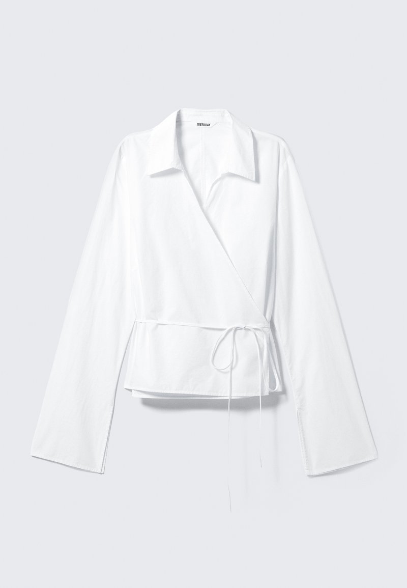 Blouse blanche à manches longues, portefeuille avec col et ceinture nouée sur le côté, posée à plat sur un fond gris clair.