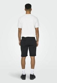 Camiseta blanca, pantalones cortos negros enrollados y zapatillas negras. La vista trasera muestra un diseño simple y un ajuste casual. Tejido texturizado, sin patrones visibles.