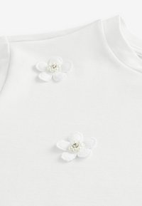 T-shirt en coton blanc avec deux fleurs décoratives brodées blanches avec des cœurs en perles sur l'épaule gauche et la zone du cou.