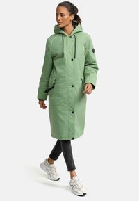 Green long coat with a hood, featuring black snap buttons and drawstrings. Worn over black pants and white sneakers; visible pockets.