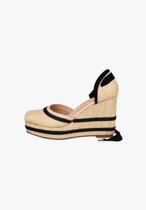 El zapato de cuña de esparto presenta un exterior similar al mimbre en beige con rayas negras, punta abierta y lazos de tela negra en el tobillo.