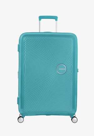 Valise rigide turquoise avec une texture striée, deux roues, une poignée rétractable et un logo circulaire à l'avant.