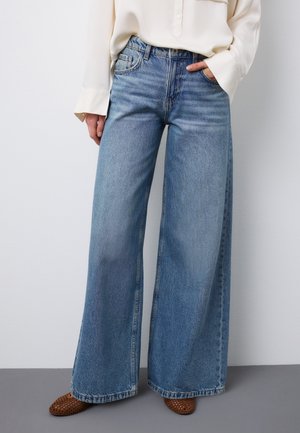 Persona che indossa jeans larghi color azzurro chiaro, una camicia bianca a maniche lunghe e scarpe intrecciate marroni, in piedi davanti a uno sfondo semplice grigio e bianco.