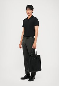 Sort læder tote bag med to robuste håndtag, der har en glat tekstur og minimalistisk design. Model iført mørkt tøj står ved siden af den.
