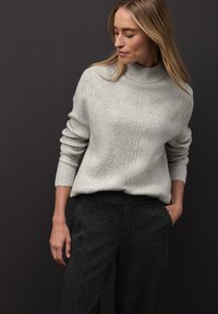 Hellgrauer, gerippter Strickpullover mit hohem Kragen und langen Ärmeln, kombiniert mit strukturierten schwarzen Hosen. Schlichtes, modernes Design.