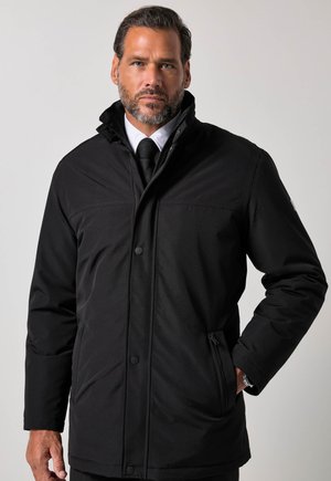 Man met baard die een zwarte winterjas draagt over een wit overhemd en een zwarte stropdas, staand voor een effen lichte achtergrond.