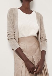 Cardigan beige con maniche lunghe, indossato sopra un maglione bianco a effetto texture, abbinato a una gonna beige a vita alta con pince e dettagli angolari.