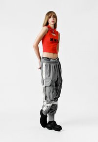 Top corto rojo con letras negras, pantalones cargo grises con cremalleras y detalles de logo, y zapatos negros texturizados. Fondo simple.
