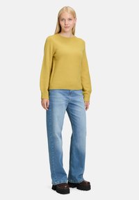 Gelbes Strickpullover mit langen Ärmeln, kombiniert mit hellblauen weit geschnittenen Jeans und dicken braunen Plateauschuhen. Studio-Setting, neutraler Hintergrund.