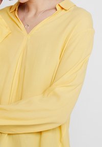 Personne portant un blouse long ample jaune à manches longues et un collier en argent avec un petit pendentif courbé, les bras croisés.