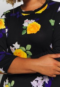 Chemise à manches longues florale avec un fond noir ; présente des roses jaunes, des fleurs blanches et des feuilles vertes ; texture douce, encolure ronde.