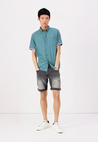 Chemise turquoise à manches courtes avec des poignets roulés, associée à un short en jean noir délavé et des baskets blanches. Design simple avec une poche poitrine.
