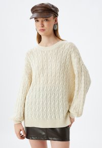Cremfarbener Strickpullover mit Zopfmuster und gerippten Bündchen. Kombination mit einem schwarzen Leder-Minirock und einer braunen Mütze.