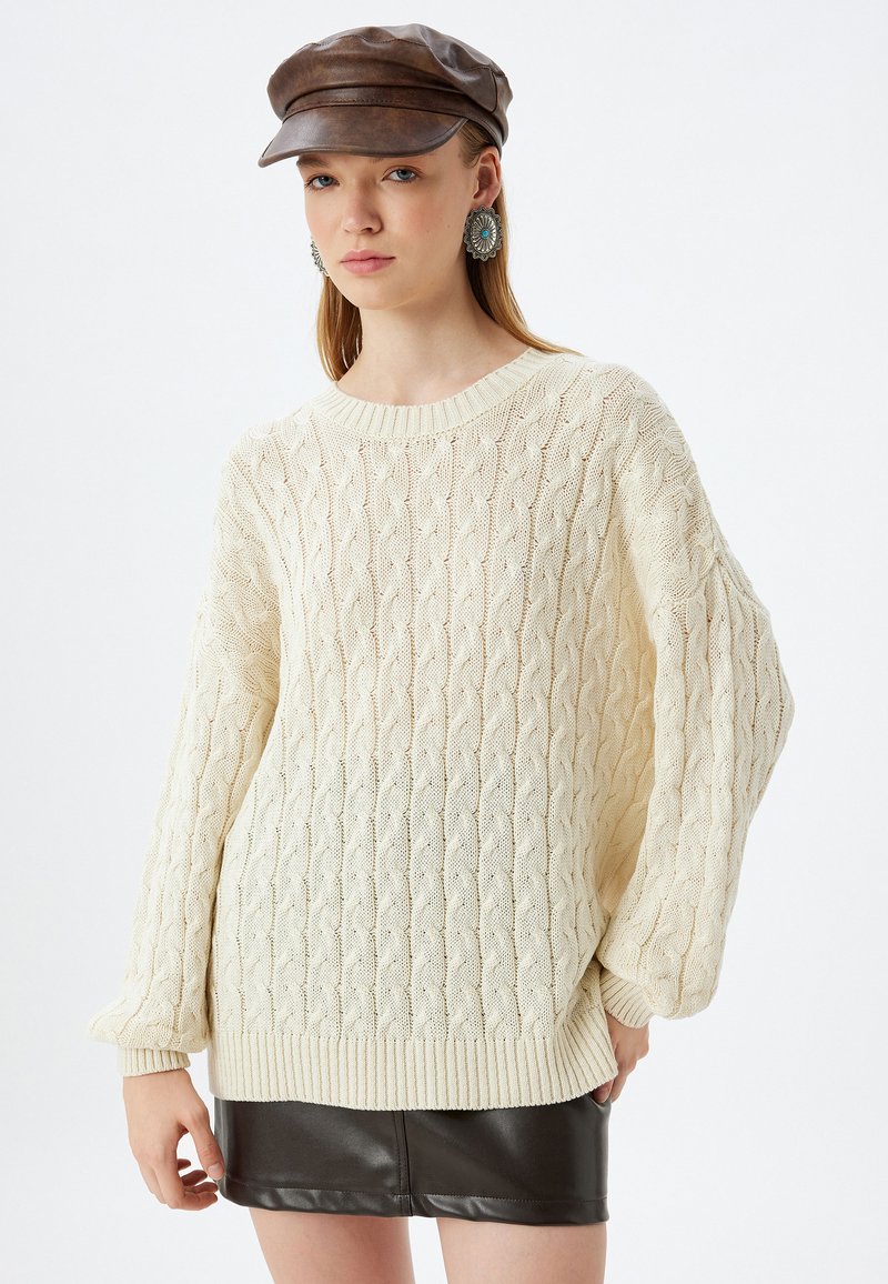 Cremfarbener Strickpullover mit Zopfmuster und gerippten Bündchen. Kombination mit einem schwarzen Leder-Minirock und einer braunen Mütze.