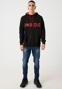 Sudadera con capucha negra y cordones rojos, con la palabra "INSIDE" en letras rojas en negrita; combinada con jeans azules y zapatillas negras.
