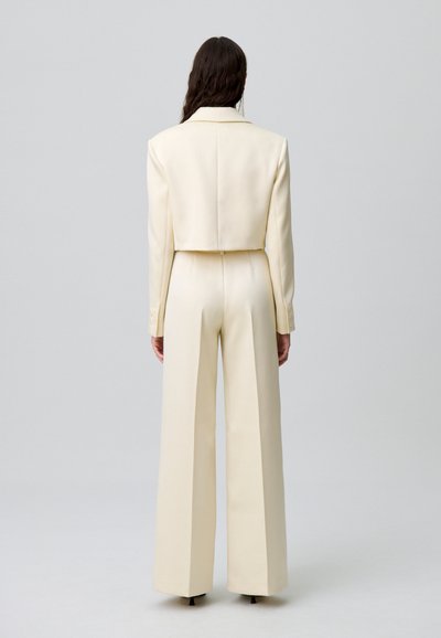 Jumpsuit ajusté crème avec une veste courte, un pantalon à jambes larges, une texture lisse et des accents de boutons aux poignets, vu de dos.