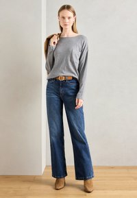 Kvinne står innendørs iført en grå langermet topp, blå vide jeans med et brunt belte, beige ankelstøvler, og holder en beige jakke over den ene skuldren.