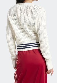 Bílý pletený svetr s otevřeným vzorem stehů, žebrovanými manžetami a lemem, obsahující námořnické pruhy na spodní části, nošený přes červenou látku.