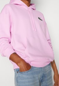 Personne portant un sweat à capuche rose clair avec un logo de crocodile sur la poitrine, les mains dans les poches d'un jean bleu clair, tatouage visible sur le poignet.