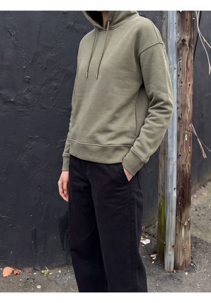 Personne portant un sweat à capuche vert olive et un pantalon noir, debout avec une main dans la poche contre un mur sombre texturé et un poteau en bois en extérieur.