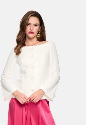 Frau mit welligem Haar, die goldene runde Ohrringe, ein weißes, langärmliges Oberteil mit Knöpfen und einen hoch taillierten, hellpinkfarbenen Rock trägt, stehend vor weißem Hintergrund.