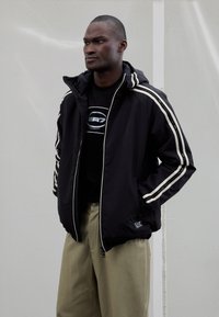 Črno jakno s kapuco z belimi črtami na rokavih, kombinirano s črno majico s kapuco z logotipom. Bež hlače. Čist dizajn in športen kroj.