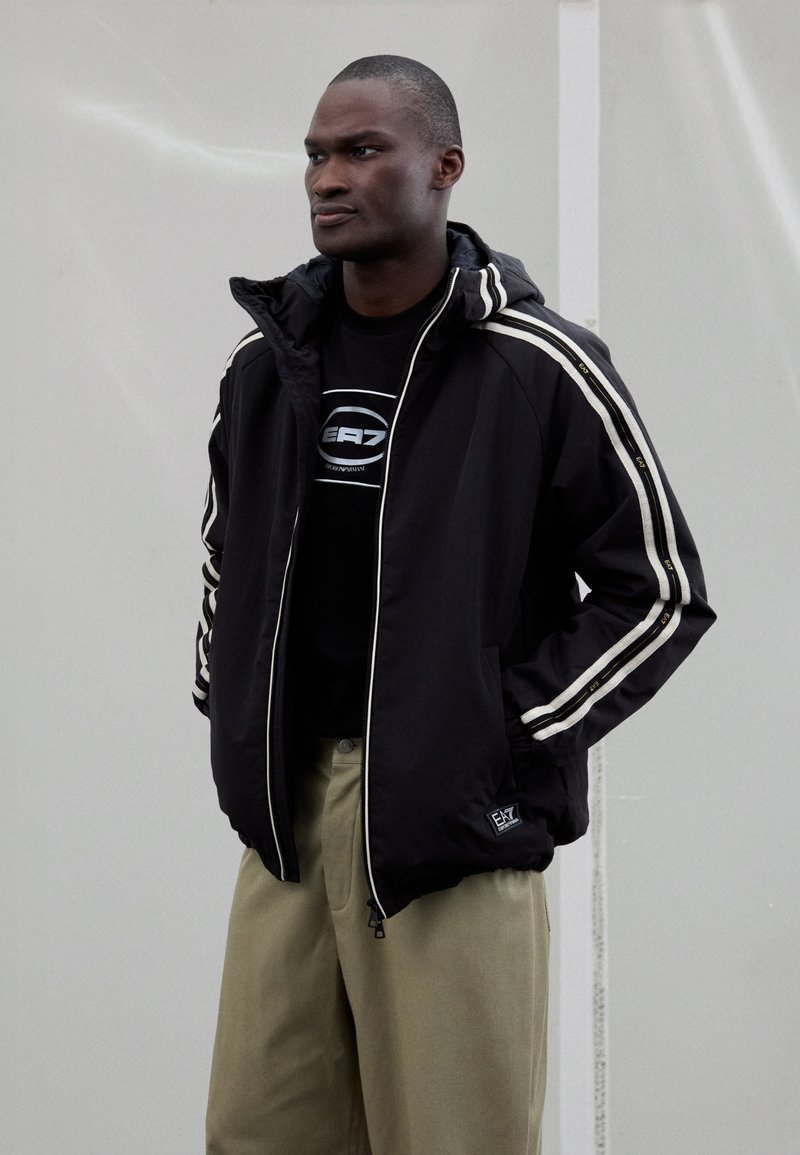 Črno jakno s kapuco z belimi črtami na rokavih, kombinirano s črno majico s kapuco z logotipom. Bež hlače. Čist dizajn in športen kroj.