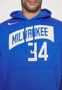 Modrá mikina s kapucí, na přední straně s bílým logem "MILWAUKEE" a číslem "34". Vyrobená z měkké látky s klokaní kapsou.