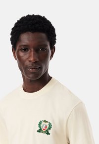 T-shirt in cotone light cream con logo Lacoste ricamato in verde e rosso, arricchito da un design a alloro sul petto. Stile classico con collo rotondo.