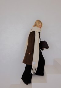 Bruine suède jas, crèmesjaal met franjes, zwarte broek met wijde pijpen en een donkere handtas. Lichte achtergrond en traptreden zorgen voor minimaal contrast.