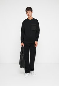 Black sweatshirt with a front pocket, paired with black loose-fit pants. The model carries a gray bag and wears white sneakers.